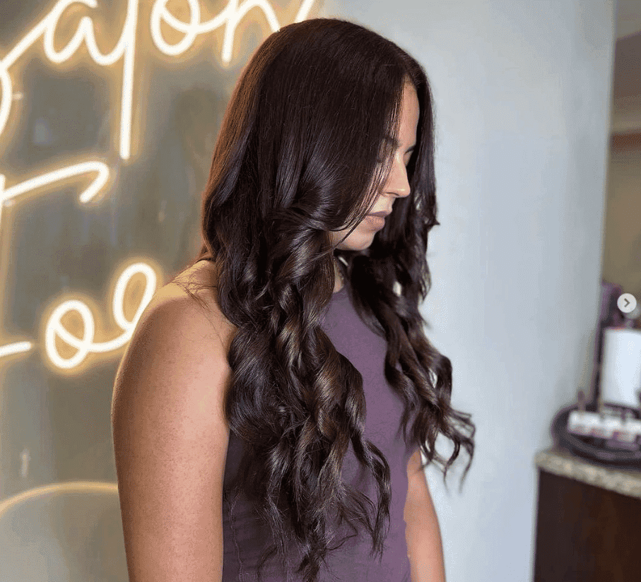 Woman with long dark hair and purple top getting her hair done in a salon with a neon sign in background.