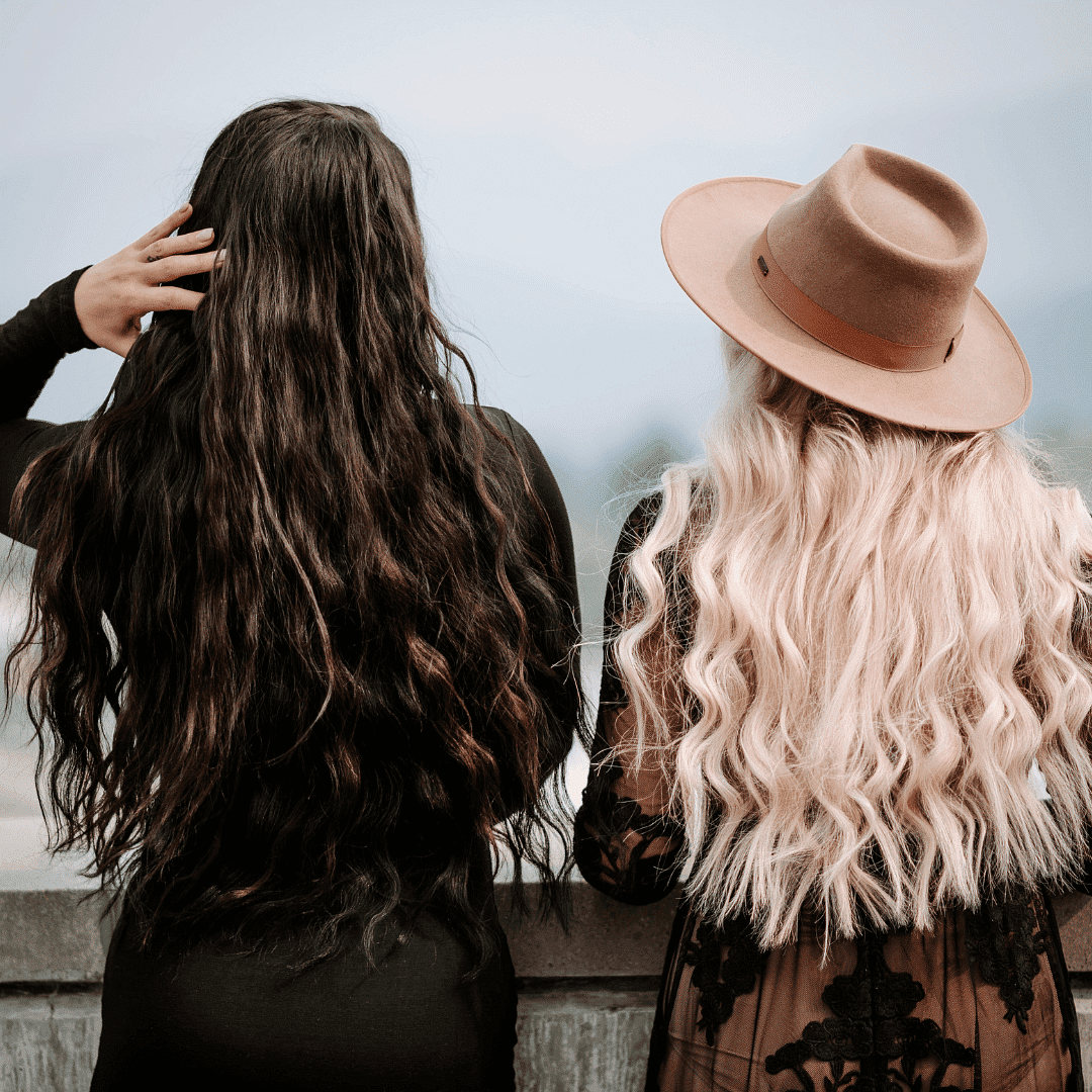 Two women with long wavy hair, one dark, one blonde, stand side by side overlooking a scenic view.